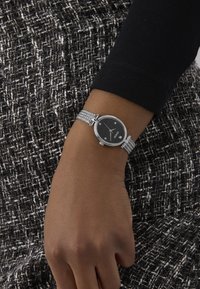 Hõbedane võrkkettaga kell musta ümmarguse kell face'iga, millel on hõbedased tunni markerid ja kuupäeva näidik, kantud randmel tekstureeritud kangaste taustal.