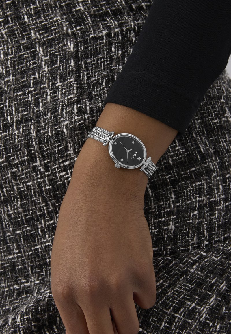 Hõbedane võrkkettaga kell musta ümmarguse kell face'iga, millel on hõbedased tunni markerid ja kuupäeva näidik, kantud randmel tekstureeritud kangaste taustal.