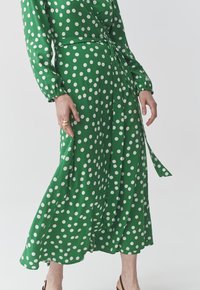 Grünes Kleid mit Punkten, langen Ärmeln, Wickeldesign, elastischen Manschetten und gebundenem Taillenband. Glattes Material mit weißen Punkten.
