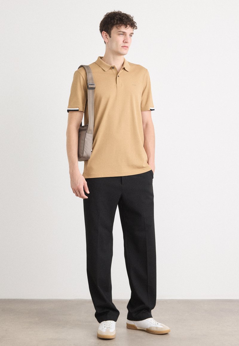 Polo shirt tan avec des poignets contrastés noir et blanc, associé à un pantalon noir et des baskets blanches, tenant un petit sac gris.