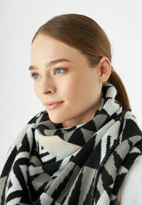 Een vrouw met blauwe ogen en bruin haar in een staart, die een grote zwarte, witte en grijze sjaal met geometrisch patroon over een witte top draagt.