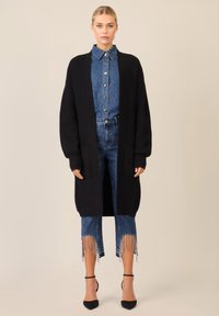 Cardigan lungo nero lavorato a maglia con maniche over size, abbinato a una camicia di jeans blu con colletto e tasche, e jeans di denim con orlo sfrangiato.