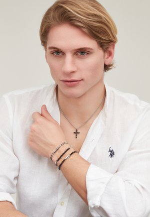 Giovane uomo con camicia bianca che indossa una collana con croce e tre braccialetti, posando con la mano vicino al collo su uno sfondo neutro.