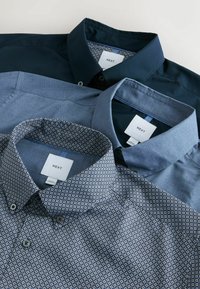 Három galléros ing fektetve: egy sötét tengerészkék, egy egyszínű világoskék és egy tengerészkék mintás, fehér geometriai mintákkal. Mindegyiken címkék vannak.