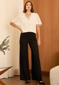 Weiße Bluse mit kurzen Ärmeln, gewelltem Ausschnitt und strukturiertem Muster, kombiniert mit hochtaillierten schwarzen weit geschnittenen Hosen. Schwarze spitze Pumps.