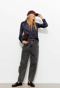 Chemise navy à motifs floraux, jeans gris larges, ceinture brune, mocassins sombres et casquette bordeaux. La tenue présente un mélange de textures.