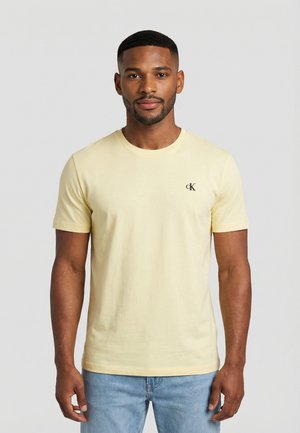 Uomo che indossa una T-shirt a maniche corte di colore giallo chiaro con un piccolo logo "cK" sul petto sinistro e jeans azzurri, in piedi davanti a uno sfondo semplice.