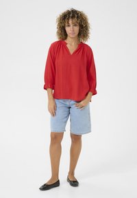 Rdeča bluzica z dolgimi rokavi in gubanjem okoli vratu v kombinaciji s svetlim denim kratkimi hlačami. Črne balerinke. Preprost, sproščen outfit.