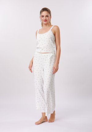 Jonge vrouw staat blootsvoets in een wit pyjamapak met kleine grijze printjes, glimlachend tegen een effen lichte achtergrond.