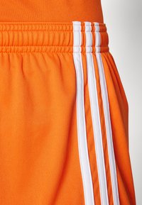 Oransje treningsshorts med teksturert stoff, med hvite vertikale striper på sidene og elastisk midje for justerbar passform.