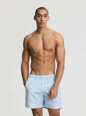 Uomo muscoloso in piedi con le mani nelle tasche, indossa pantaloncini da bagno azzurri con piccole stampe di aragoste arancioni su sfondo uniforme.