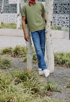 Person trägt grünes T-Shirt, blaue Jeans, weiße Turnschuhe und rotes Bandana, lehnt zwischen zwei weißen Baumstämmen in einem städtischen Gartenbereich.