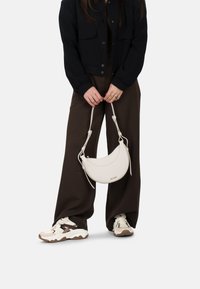 Sac en cuir blanc en forme de croissant avec une fine bandoulière, porté par une personne vêtue d'une veste noire et de pantalons larges marron, associés à des baskets volumineuses.