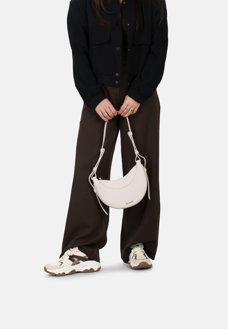 Bolso de cuero blanco en forma de media luna con una banda delgada, sostenido por una persona que lleva una chaqueta negra y pantalones anchos marrones, combinados con zapatillas chunky.