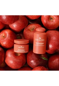 Fondo temático de manzana roja con dos productos de ORIGINS: un frasco pequeño y una botella alta, ambos en un color coral-naranja a juego con texto blanco.