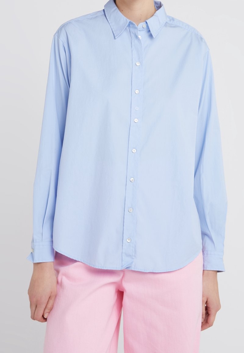 Ljusblå långärmad skjorta med knappar och spetsig krage, rundad nederkant och subtil glans; kombinerad med rosa byxor.