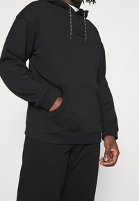 Black hooded sweatshirt with a front pocket, textured fabric, contrasting drawstring, and a woven label on the chest.