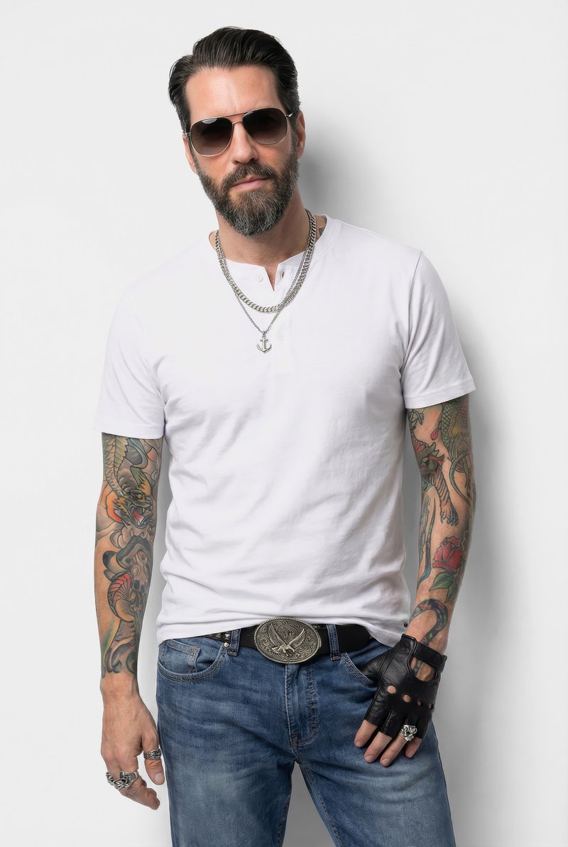 Homme avec barbe et lunettes de soleil portant un t-shirt blanc, un jean bleu, un gant en cuir, des chaînes et des tatouages colorés détaillés sur les bras, debout contre un mur blanc.