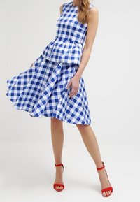 Frau in einem blau-weißen karierten ärmellosen Kleid mit ausgestellt Rock, kombiniert mit roten High-Heels mit Knöchelriemen.