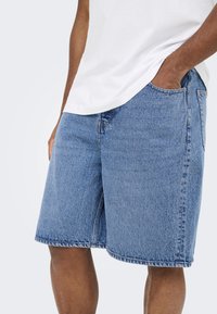 Mand iført lyseblå denimshorts og en hvid t-shirt med den ene hånd i shorts-lommen mod en ensfarvet baggrund.