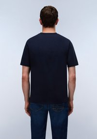 Mężczyzna stoi tyłem, ubrany w gładki ciemnogranatowy t-shirt i niebieskie dżinsy na jasnoszarym tle.