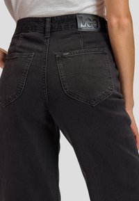 Back view of person wearing high-waisted black Lee jeans with visible back pockets and leather brand patch on waistband.