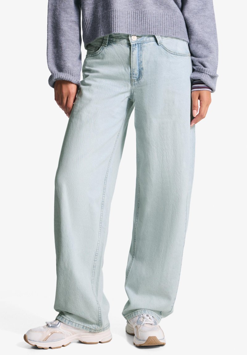 Svijetloplavi široki traperice od denima, s dugmetom zatvaranjem, prednjim džepovima i suptilnom teksturom, uparenih s bijelim tenisicama.