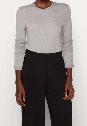 Person iført en lysegrå langærmet skjorte, der er stoppet ned i højtaljede sorte plisserede bukser, stående mod en ensfarvet baggrund.