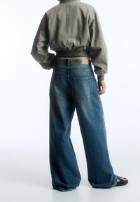 Cazadora corta de color verde oliva con mangas abullonadas, combinada con vaqueros de mezclilla azul lavado medio de pierna ancha con dos bolsillos traseros.