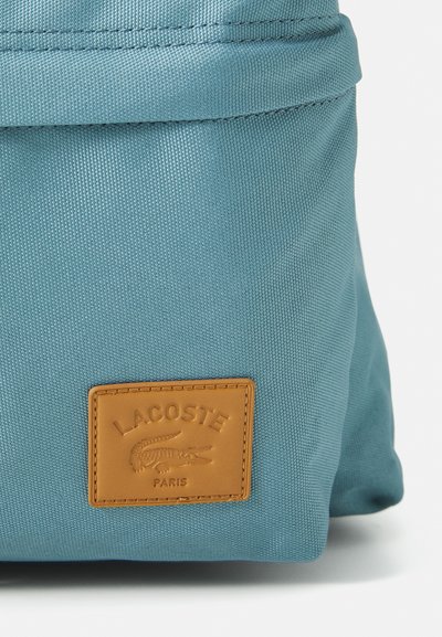 Sac à dos en toile bleue avec une surface texturée, comportant une étiquette en cuir beige embossée avec "LACOSTE PARIS" et un logo de crocodile.