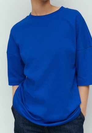 Osoba ubrana w jasnoniebieską, luźną koszulkę z krótkim rękawem i okrągłym dekoltem oraz ciemnoniebieskie jeansy na jednolitym tle.