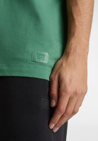 Chemise verte à manches courtes avec une texture lisse, dotée d'un patch logo Vans rectangulaire à l'ourlet, assortie à un pantalon noir.