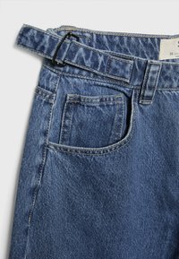 Jean en denim d'un bleu moyen, avec une ceinture à boucle noire, un détail de poche et des surpiqûres contrastées sur une surface texturée.