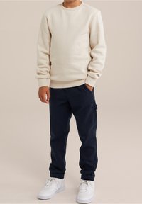 Ljus beige sweatshirt med ribbad mudd och nederkant, matchad med mörkblå byxor som har sidofickor. Vita sneakers fulländar outfiten.