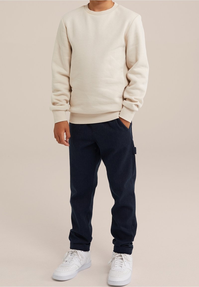 Ljus beige sweatshirt med ribbad mudd och nederkant, matchad med mörkblå byxor som har sidofickor. Vita sneakers fulländar outfiten.