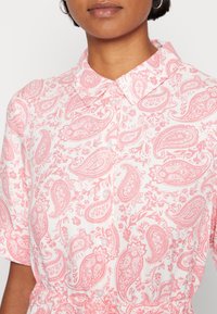 Camisa de estampado de paisley rosa con cuello, mangas cortas y cierre de botones. La tela presenta acentos florales y detalles texturizados.