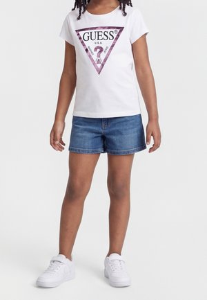 Barn iført hvid Guess t-shirt med pink trekantet logo, blå denimshorts og hvide sneakers, stående mod ensfarvet baggrund.