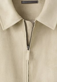 Veste beige avec col et fermeture éclair en métal argenté partiellement zippée, dotée d'une languette de tirage en cuir et d'une étiquette de marque intérieure indiquant MNG.