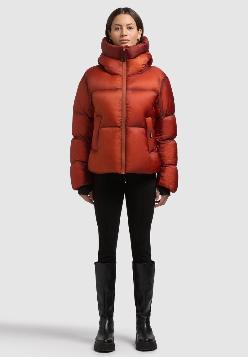 Puchowa czerwona kurtka z wysokim kołnierzem, zamkiem z przodu i dwoma bocznymi kieszeniami. Całość dopełniają czarne spodnie i czarne kozaki za kolano.