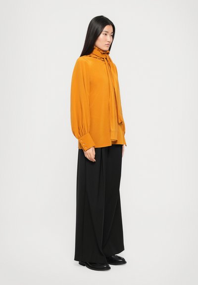 Mosterdgele blouse met lange mouwen en strikdetail, gecombineerd met losse zwarte broeken en platte zwarte schoenen. Zachte textuur, minimalistisch ontwerp.