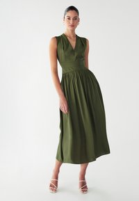 Maslinasto zelena haljina bez rukava s V-izrezom, prilagođenim tijelom i nabranom suknjom. Uparena s bijelim sandalama s remenčićima.