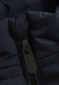 Veste matelassée noire en tissu avec fermeture éclair et boutons-pression. Comprend des panneaux cousus et une texture lisse. Logo sur la tirette de la fermeture éclair.