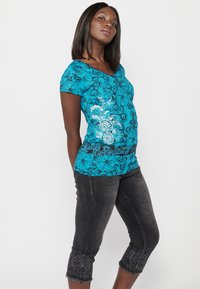 Haut à motifs floraux teal avec des manches courtes et un décolleté en V. Assorti à un pantalon court gris foncé avec des ourlets décoratifs. Texture de tissu lisse.