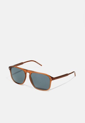 Occhiali da sole con montatura in plastica marrone e lenti quadrate, dotati di vetro scuro. Astine sottili in marrone con dettagli sottili.