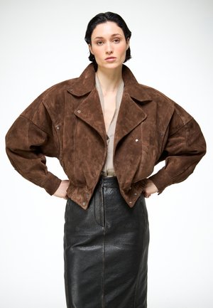 Vrouw met kort donker haar, gekleed in een oversized bruine suède jas en een hooggetailleerde zwarte leren rok, staande met handen in de zij.