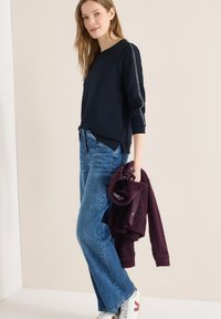 Femme portant un haut à manches longues bleu marine avec une bande latérale, un jean bleu et des baskets blanches, tenant une veste bordeaux pliée dans sa main.