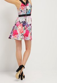 Robe fleurie avec des roses multicolores et des accents foncés, design sans manches, corsage ajusté, jupe évasée, associée à des chaussures noires à talons hauts.