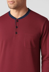 Maglietta henley a maniche lunghe color bordeaux con colletto a coste blu navy, tre bottoni neri sulla patta e tasca sul petto sinistro. Texture liscia.
