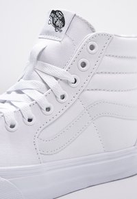 Basket Vans blanche montante avec lacets, œillets métalliques, bande latérale emblématique et logo "Vans Off The Wall" sur la languette.