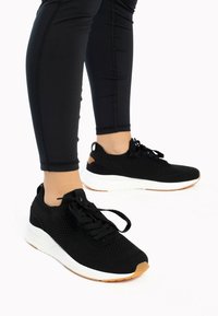 Black athletic sneakers with a mesh upper, white sole, and brown accent on the heel. Black laces and elastic collar for easy wear.
